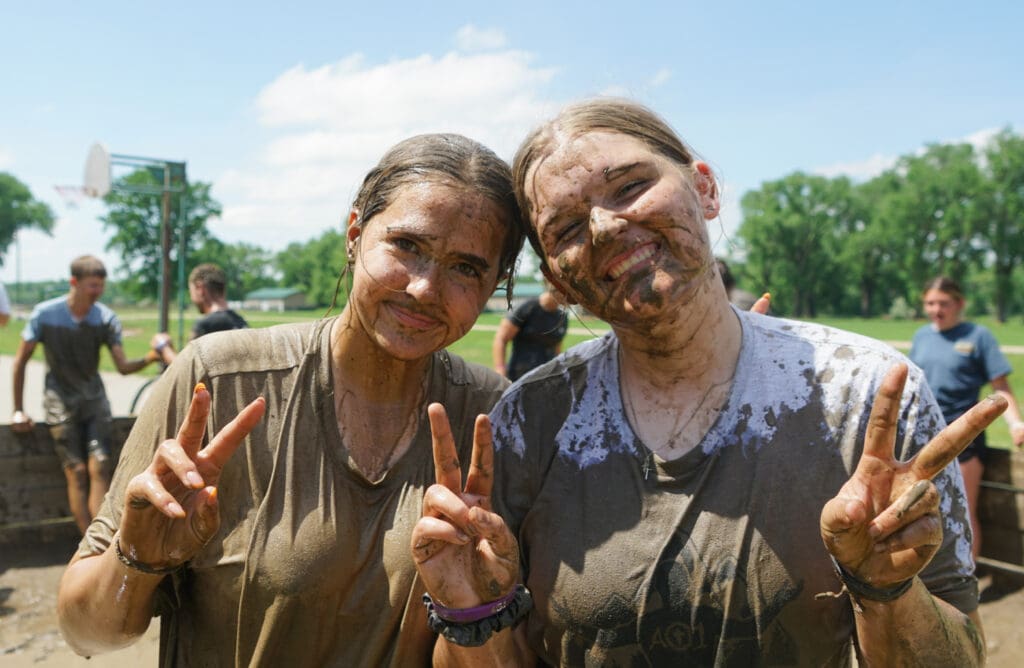 Two young women, covered in mud, smile and make peace signs after completing an obstacle course at the AO1 Foundation's Camp Conquerors.