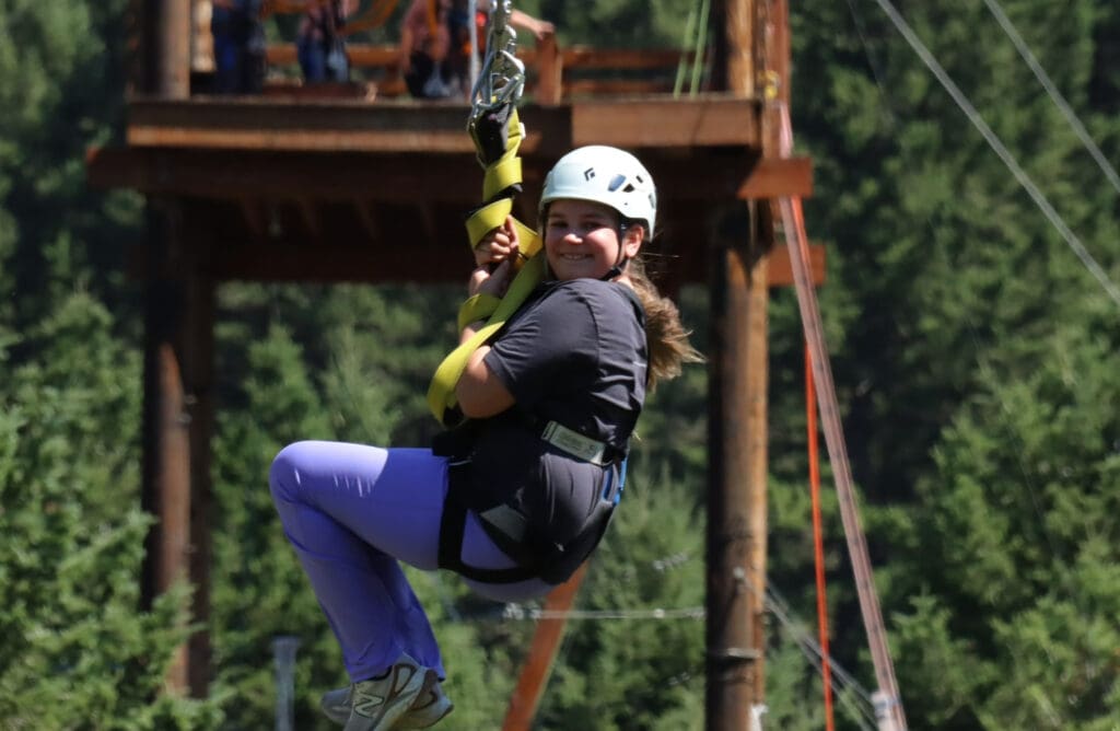 A young girl with a helmet smiles while riding a zipline at the AO1 Foundation's Mountain Movers event, with a forest and wooden platform in the background.