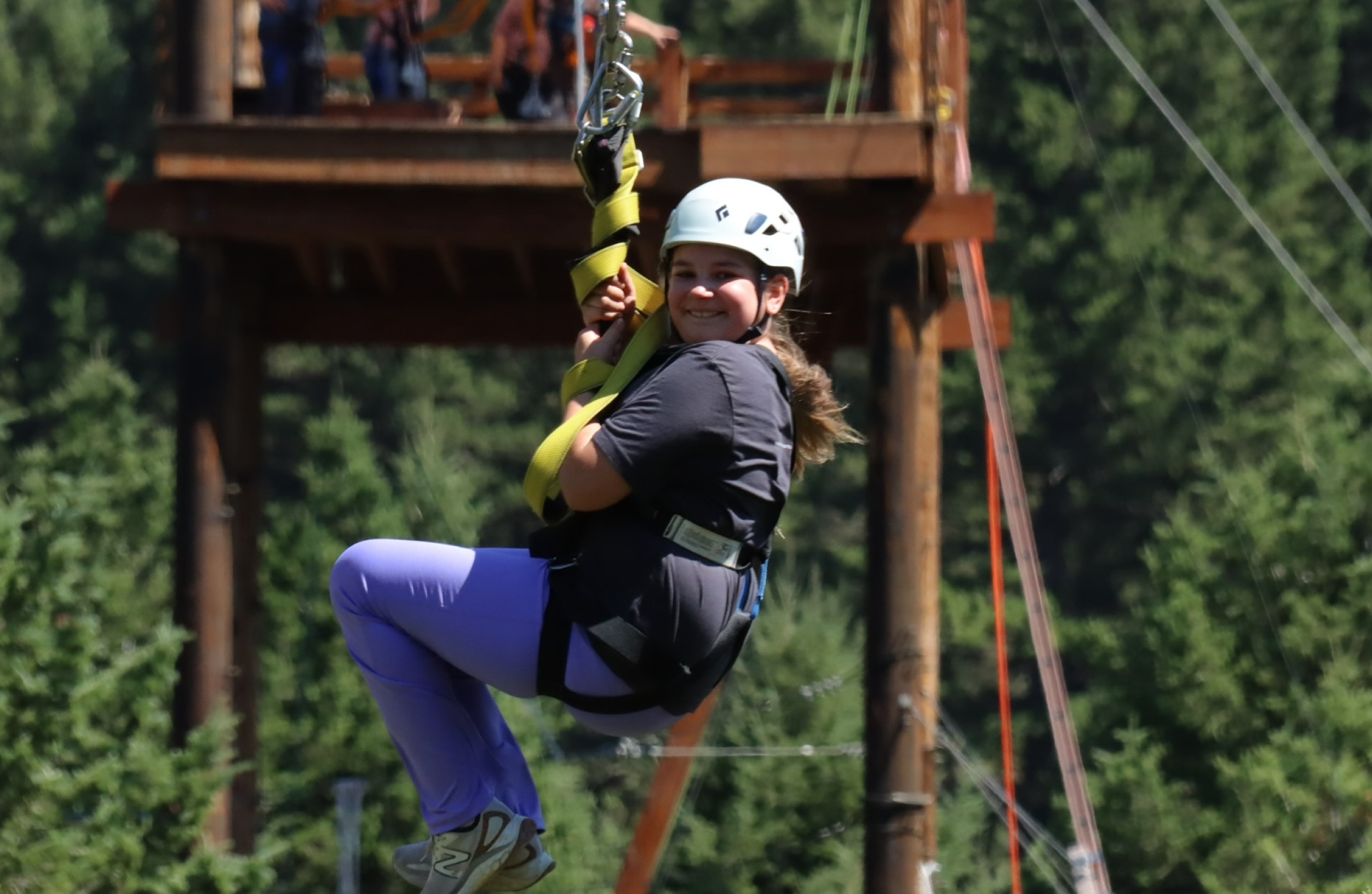 A young girl with a helmet smiles while riding a zipline at the AO1 Foundation's Mountain Movers event, with a forest and wooden platform in the background.